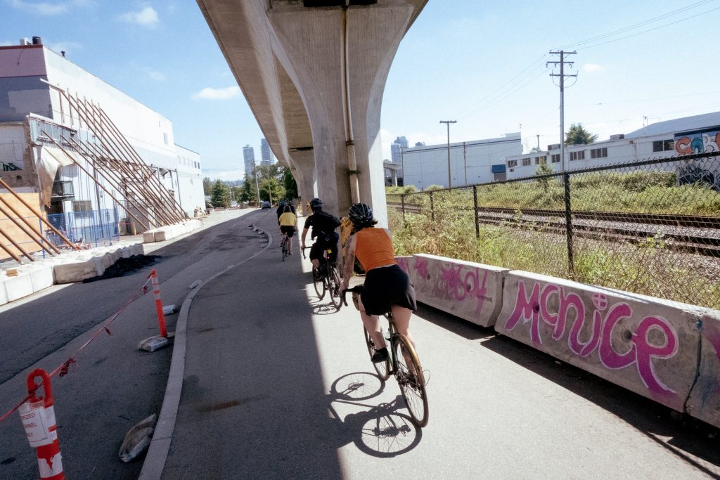 A group of cyclists ride underneath a bridge on a sunny day. There is graffiti on the cement barrier beside them.