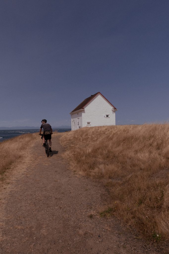 A cyclist rides alongside the ocean toward a small white house that sits at the edge of the ocean.