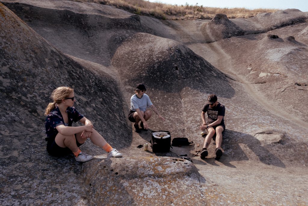 Three campers sit by a cooler on a rock face by the ocean.