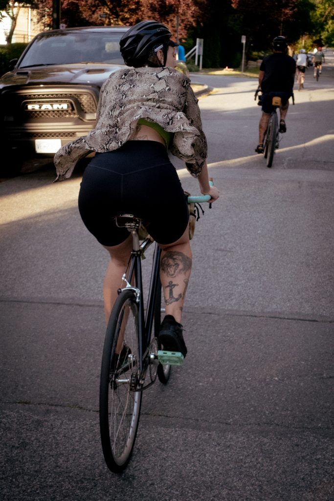 A woman rides a bike down a street in a group of riders with her shirt flapping in the wind.