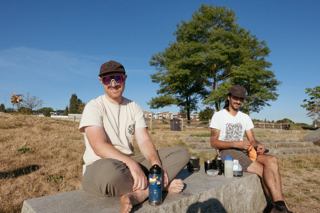 Two cyclits sit on a stone bench in front of the ocean. One is wearing sunglasses and smiling at the camera, while the other is brewing coffee.