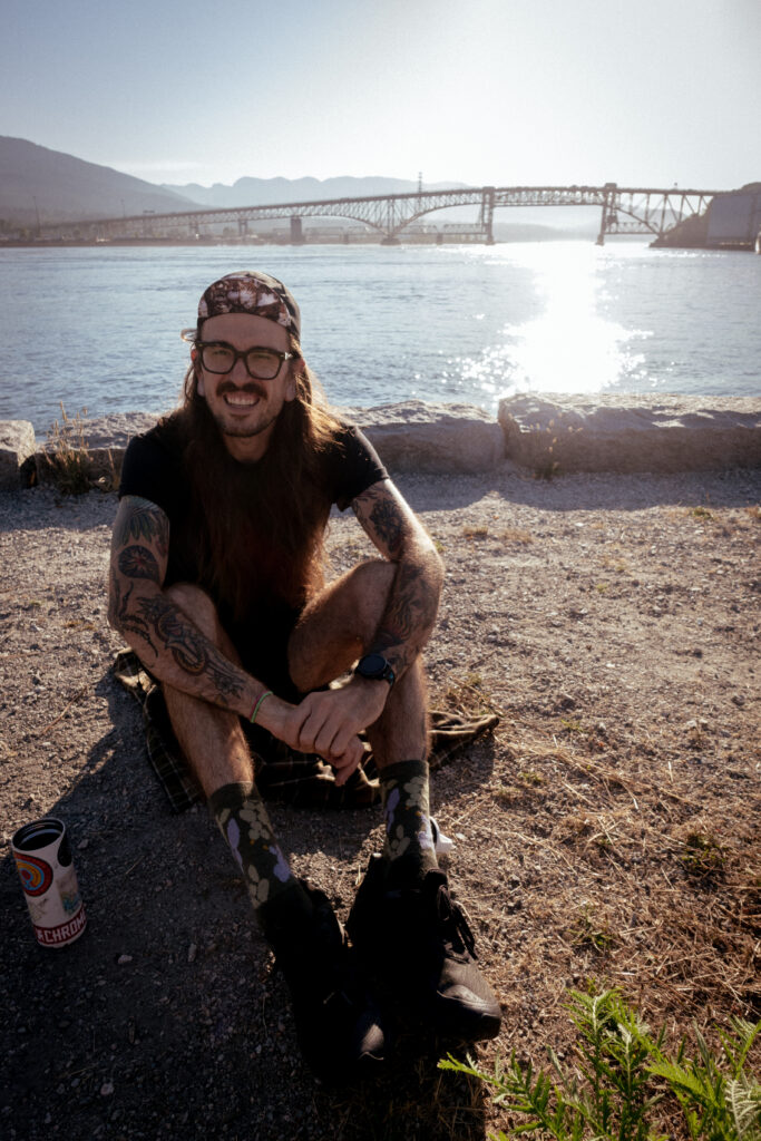 A cyclist sits on the ground with a coffee cup in front of him with the Iron Workers Bridge behind him.