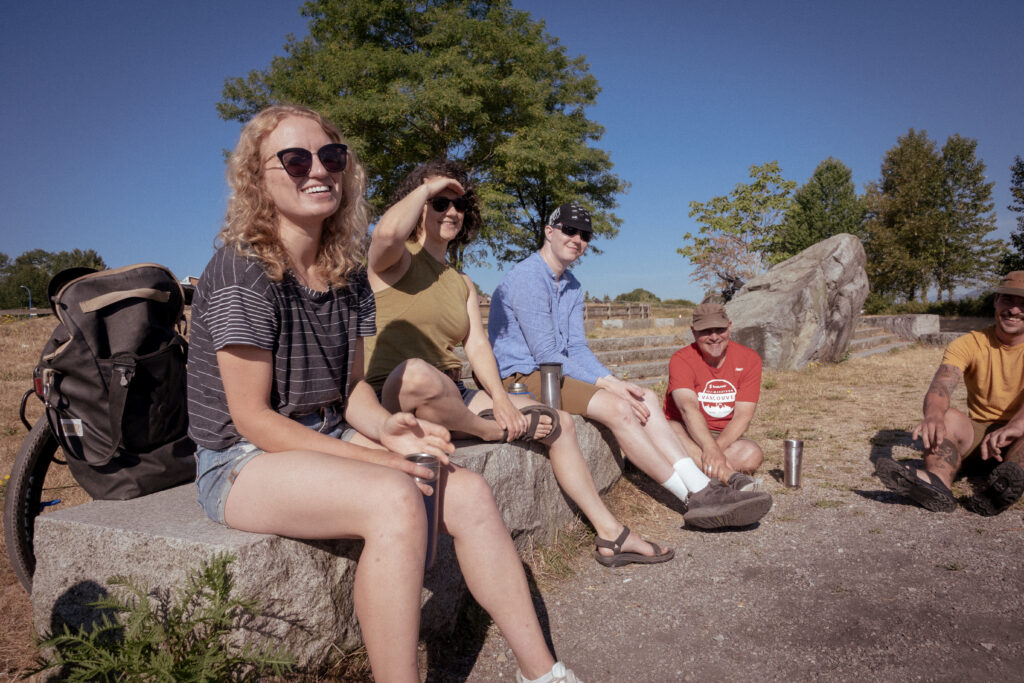 A group of 5 cyclists look at the camera smiling a drinking coffee together at an oceanfront park. Some sit on a stone bench, while one is on the group.