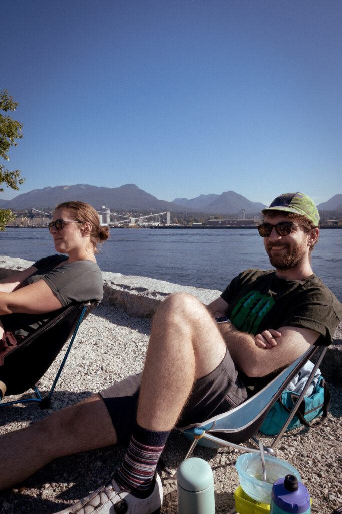 Two cyclists sit in camp chairs smiling and brewing coffee at a local meet-up event