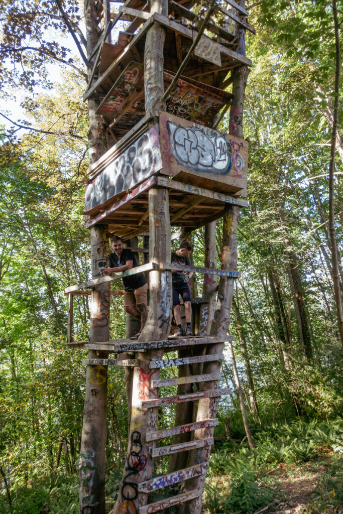 A large 3 or four-story treehouse is hidden in the forest with two people on the first story. The two people are smiling looking outwards. The treefort is spray painted with graffiti.