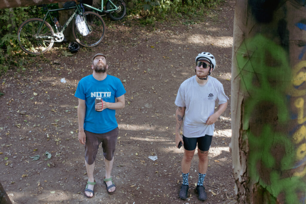 Two cyclists look upwards from the ground towards a hidden treefort in the forest. One is drinking a beer.