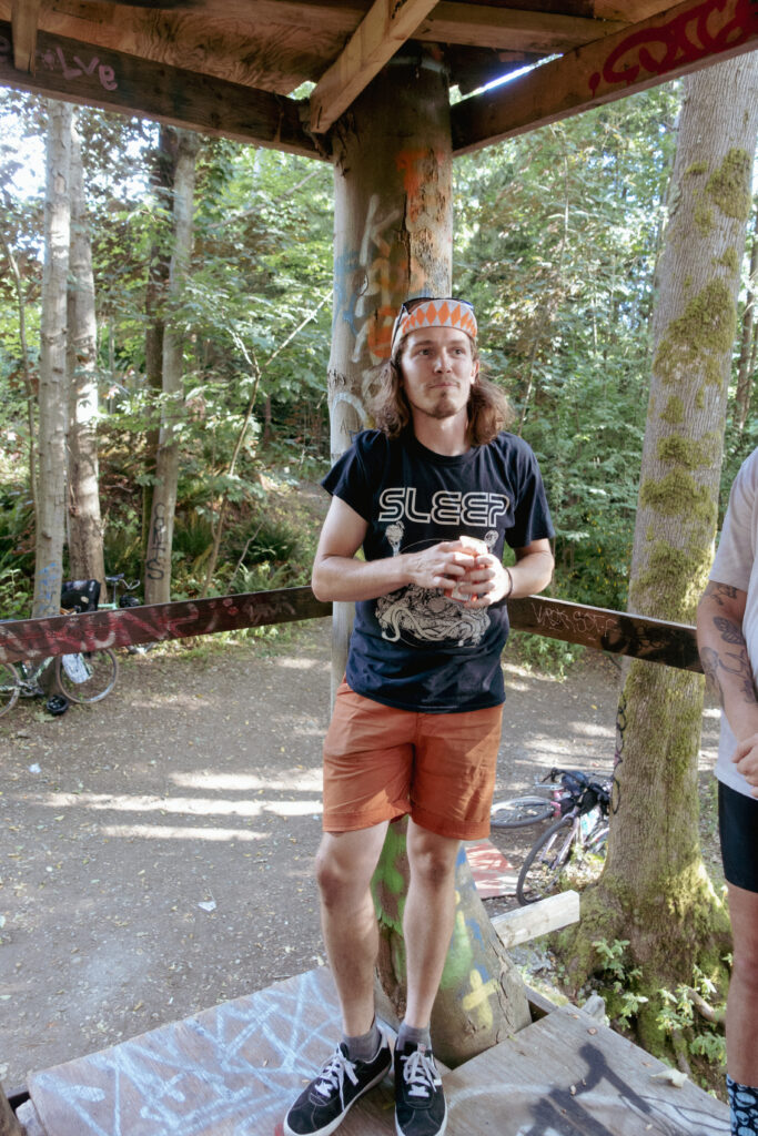 A man with a cyclist's cap and a band t-shirt is a drinking a beer in a tree fort that is spray painted with grafetti