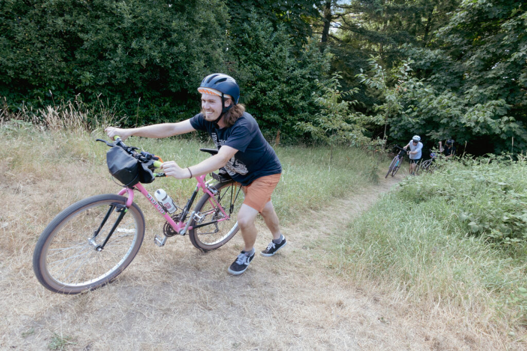 A smiling cyclist pushes a bike up hill coming out from the forest. The bike is an older 90s mountain bike style