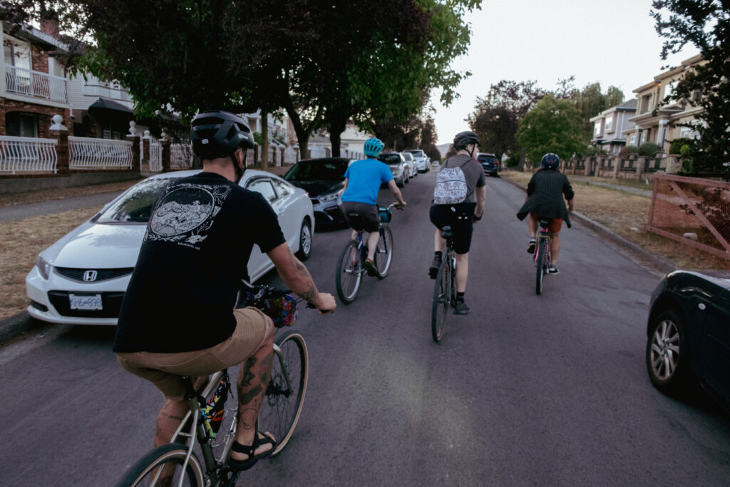 A group of 4 cyclists ride down a street. The one closest to the camera is riding in sandles.