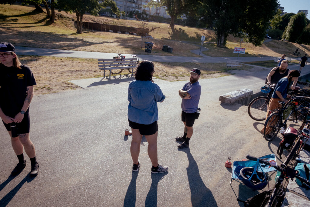 A group of cyclists chat with deep shadows casting on them. Two are talking in the center of the frame, a man and a woman.