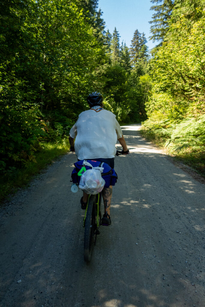 A cyclist rides their bike on a forest service road with a bag of cans hanging off of the back